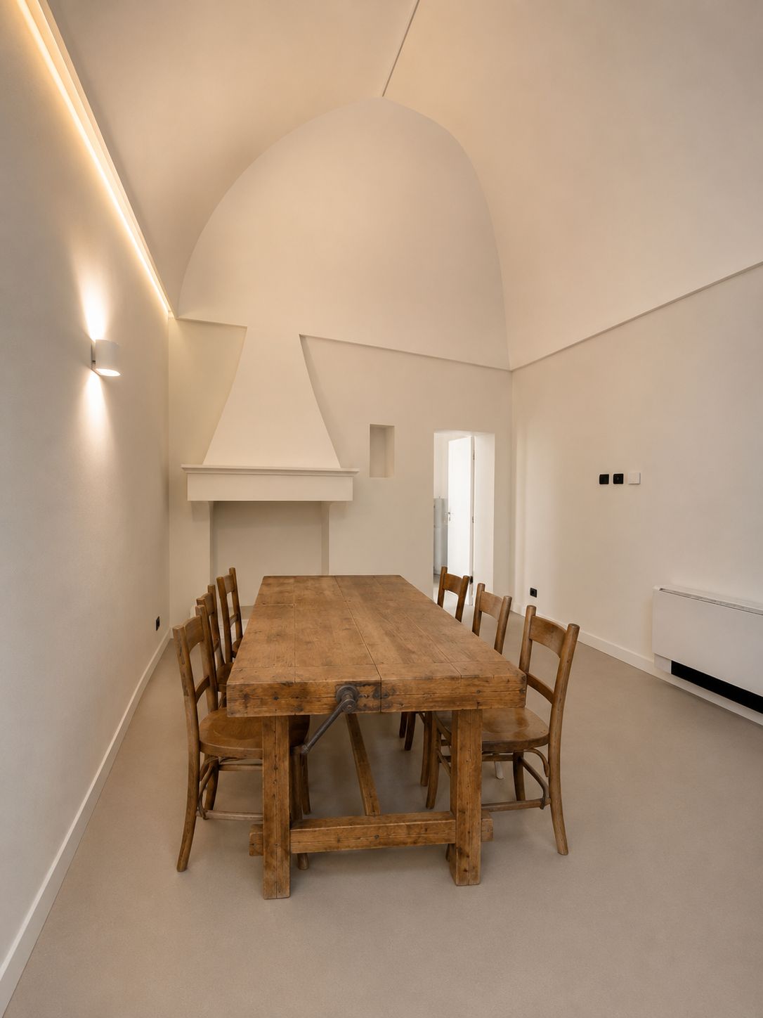 The dining room of Casa Andrea in use — an antique carpenter's workbench found at a brocante, now the dining table, with chairs under the white dome vault, the conical plaster hearth beside it, microcement floor, Puglia.