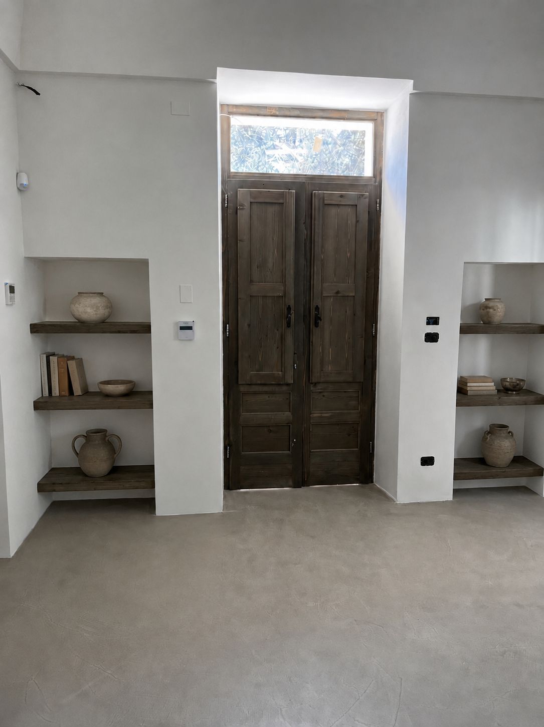 The entrance door seen from the dining room — a restored antique wooden double door with a painted majolica transom, flanked by two built-in niches with wooden shelves holding terracotta jars, microcement floor, Puglia.