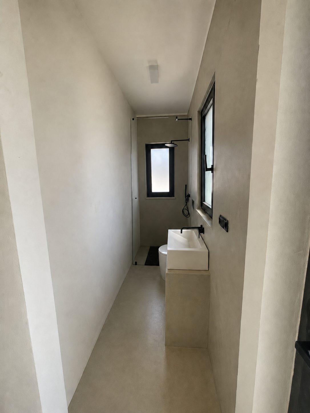 The guest bathroom of Casa Andrea — a narrow microcement room in a warm tone, a built-in washbasin with a black wall-mounted tap, a black-framed window, a walk-in shower at the far end, Puglia.