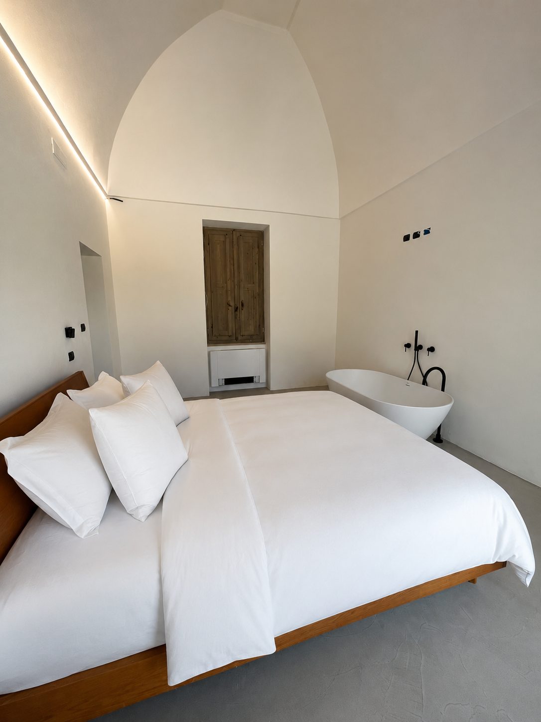 The master bedroom of Casa Andrea under its vault — wooden bed and headboard, white linen, a wooden-shuttered window recess in the wall, a freestanding sculptural tub set in the room with a black wall-mounted tap, indirect cove lighting, Puglia.
