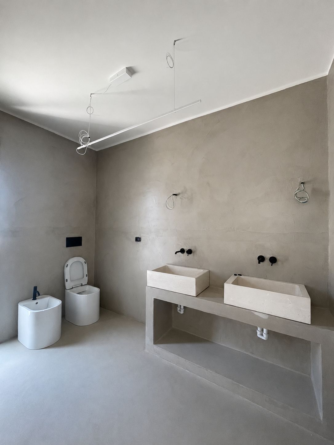 The master en-suite bathroom of Casa Andrea — a microcement double vanity with two basins and black wall-mounted taps, a bidet and WC, walls and floor in one warm tone, Puglia.