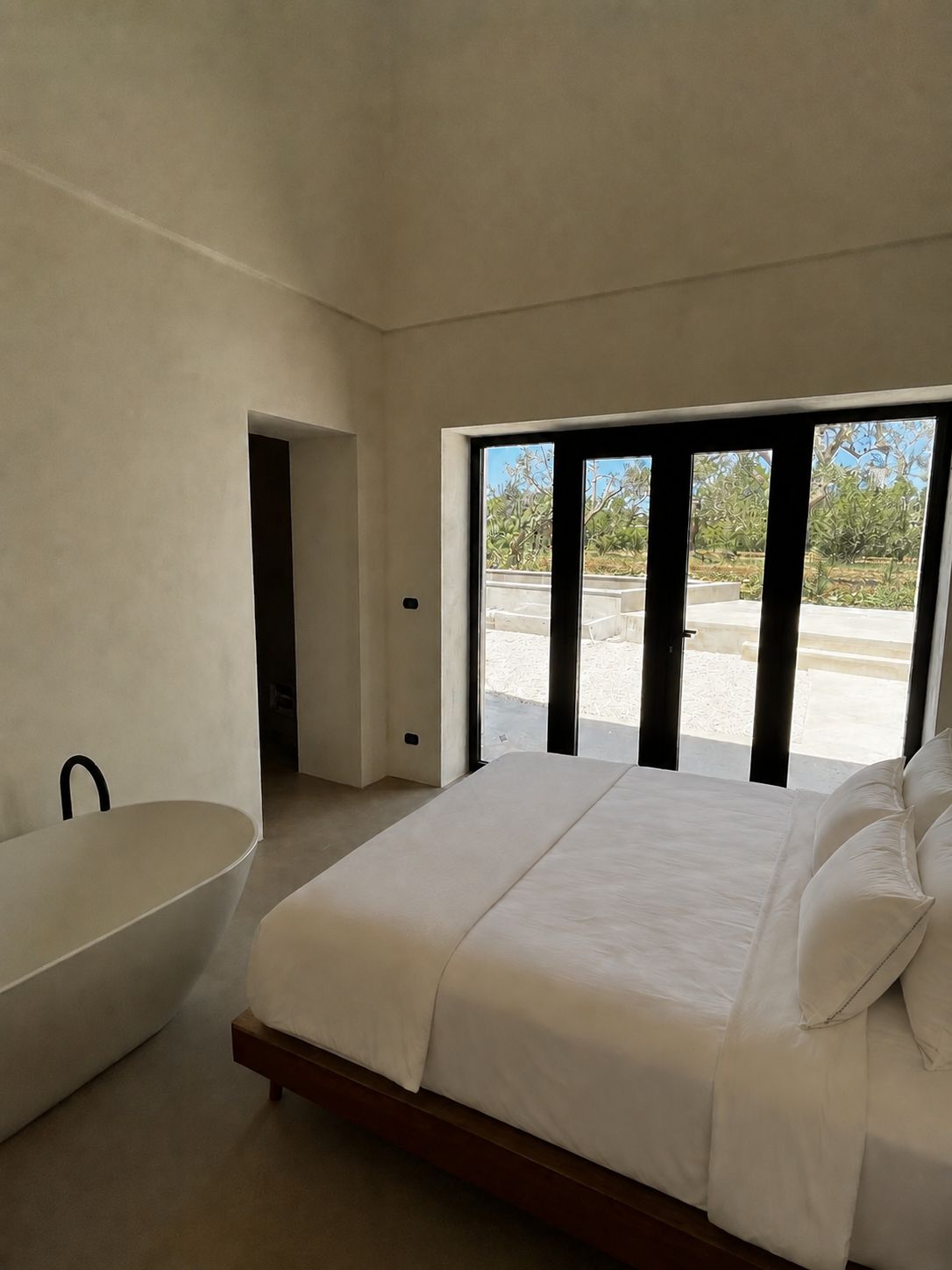 The master bedroom from another angle — a freestanding sculptural tub by the bed, full-height black-framed glazed doors opening onto the olive grove, microcement floor, Puglia.