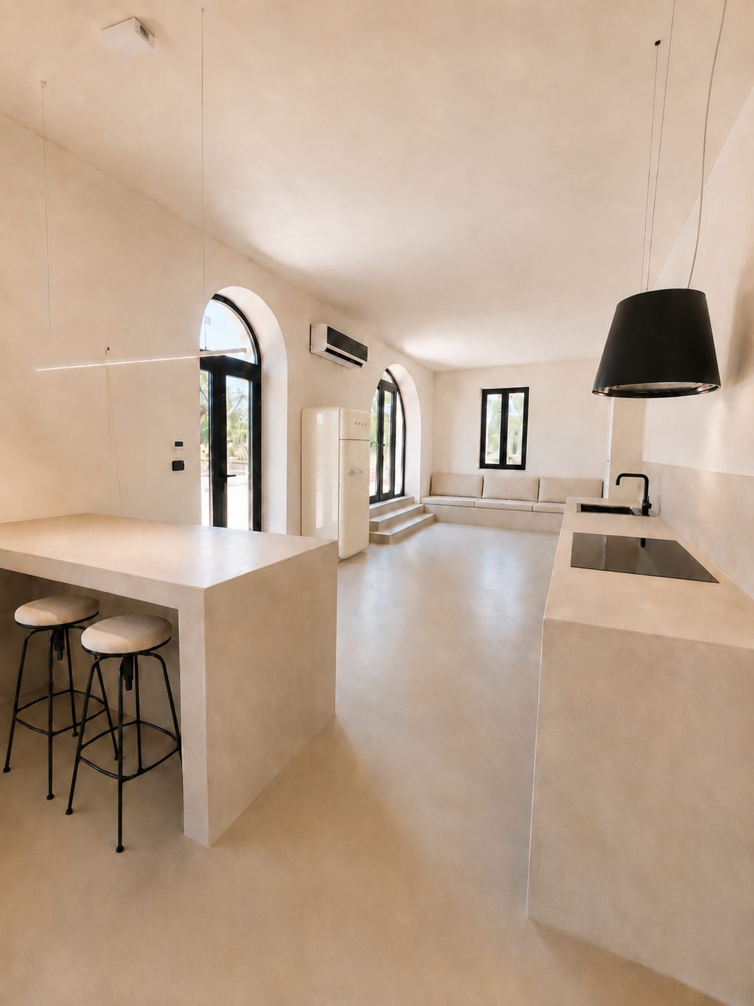 The open living room and kitchen of Casa Andrea — a stone bar peninsula with an open shelf for stools to come, the kitchen counter along the wall, two arched glass windows onto the courtyard, a black pendant lamp, microcement floor, Puglia.