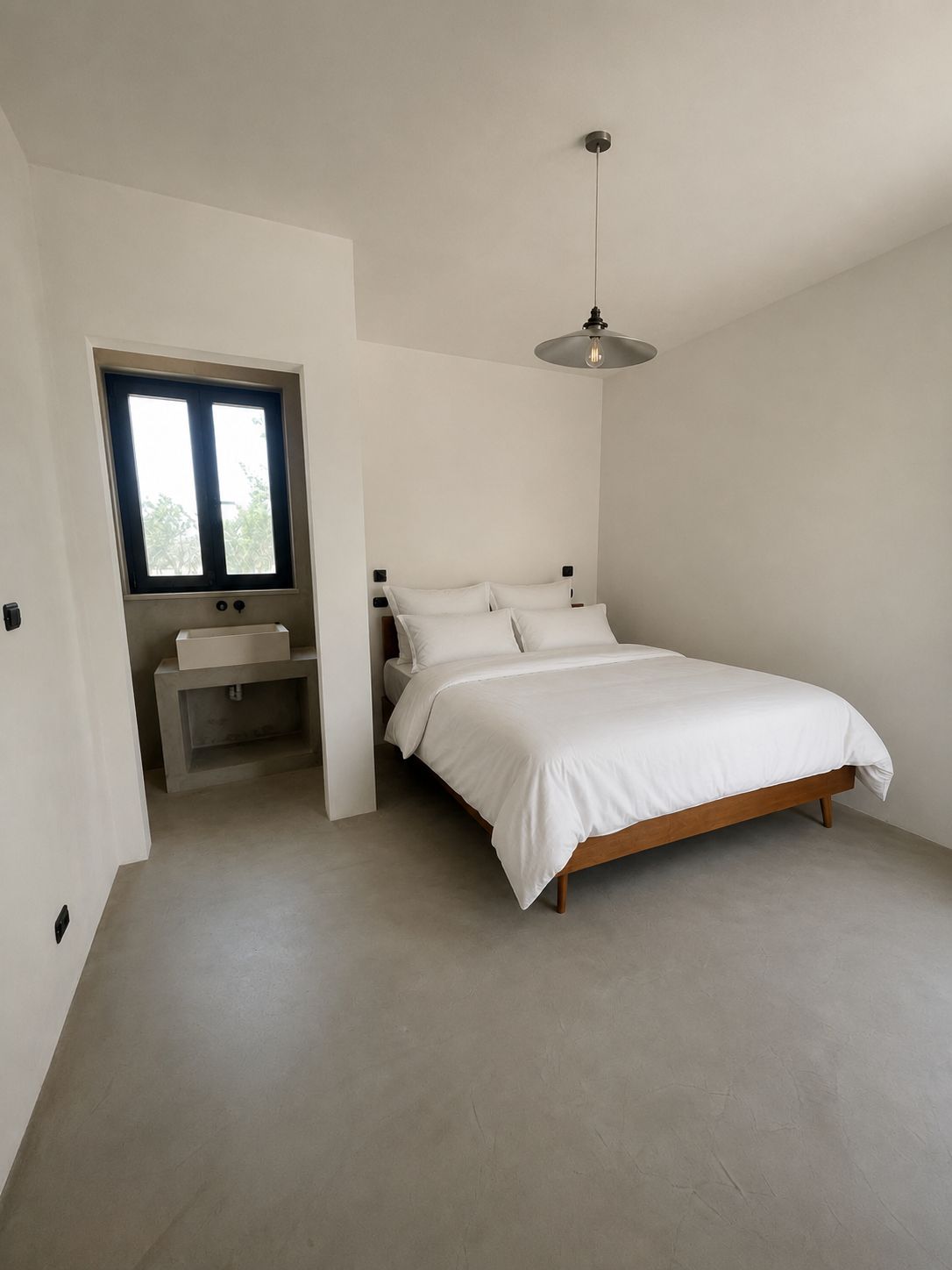The third en-suite bedroom at Casa Andrea — microcement walls and floor in a warm tone, a wooden bed with white linen, an opening to the en-suite with its washbasin, Puglia.