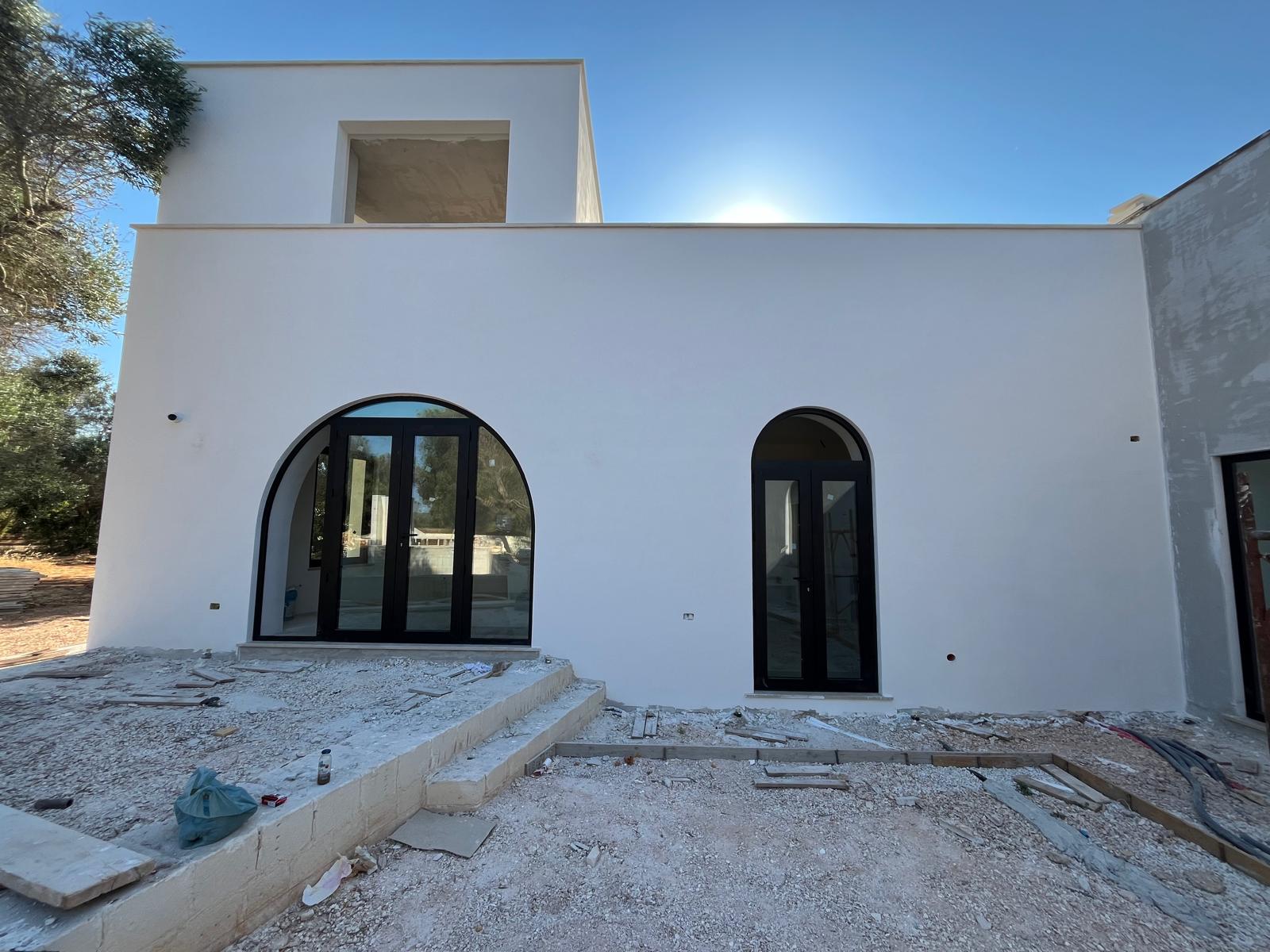 The new Living Wing of Casa Andrea — two-storey white intonaco facade with two arched openings on the ground floor and the rooftop bureau visible above, a centenary olive on the left, Puglia.