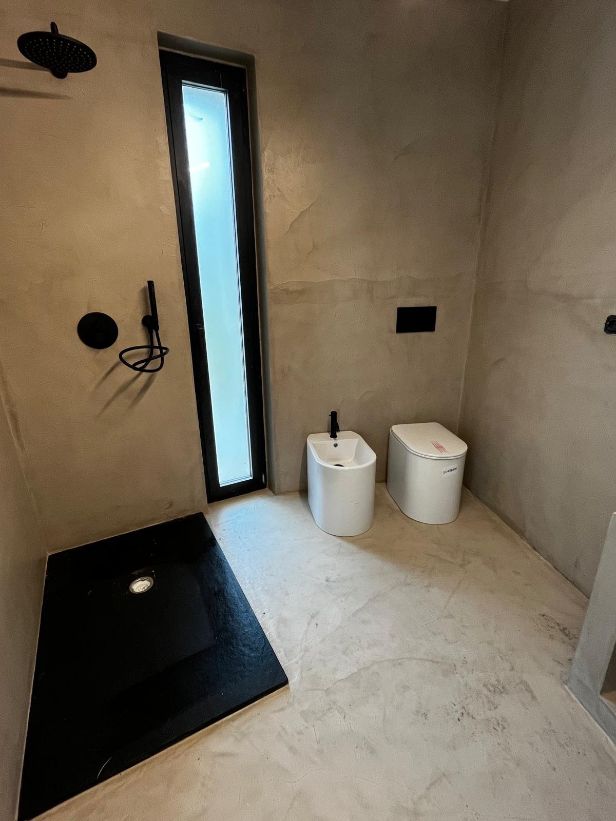 An en-suite bathroom at Casa Andrea with a tadelakt walk-in shower — warm-grey lime walls, hand-trowelled finish, brass tap, terracotta floor, Puglia.
