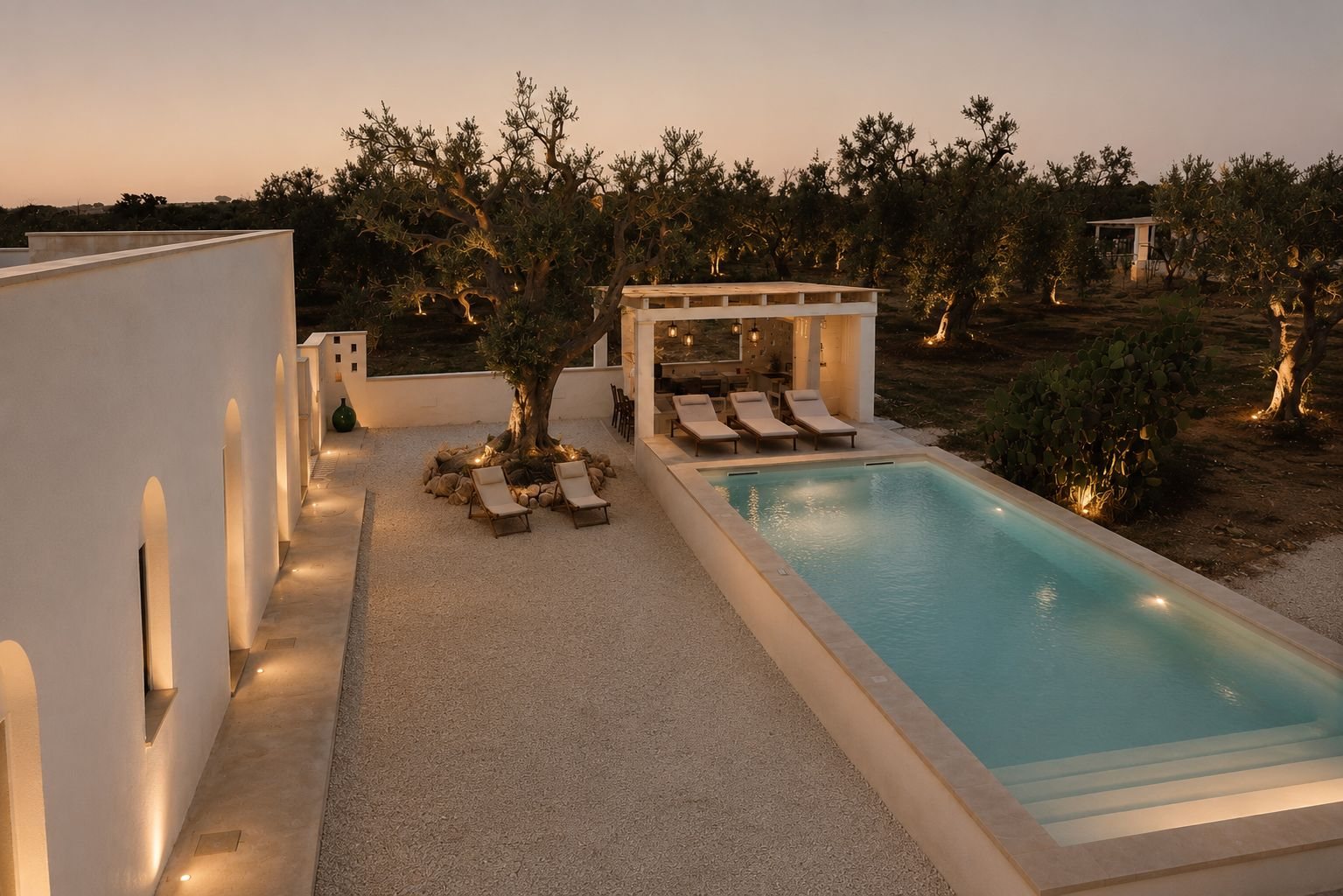 Casa Andrea at dusk — the lit pool, the centenary olive and the summer kitchen under its pergola, the grove beyond, Puglia.